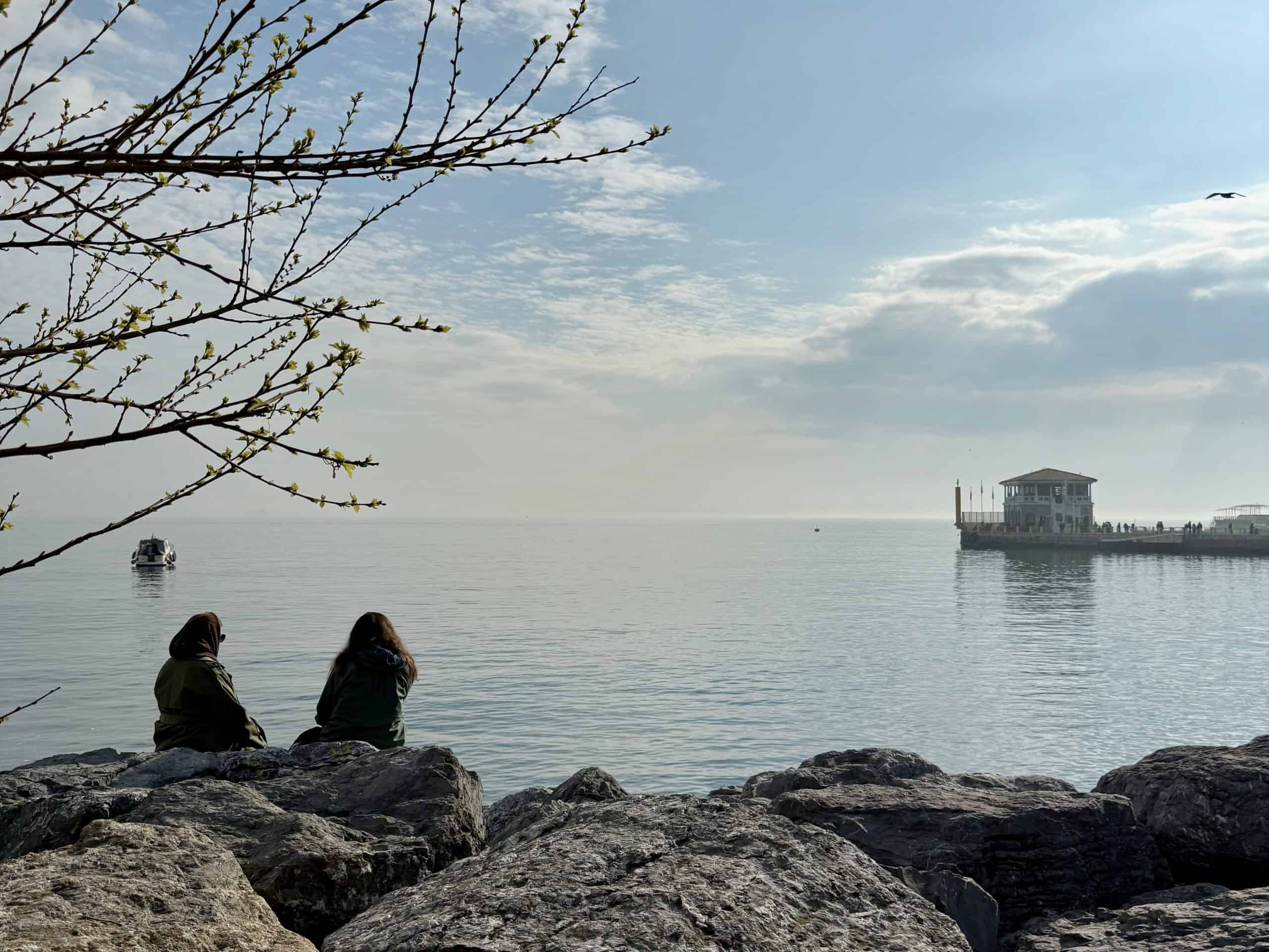 Two women sitting on rocks besides the sea looking out towards the Moda Pier