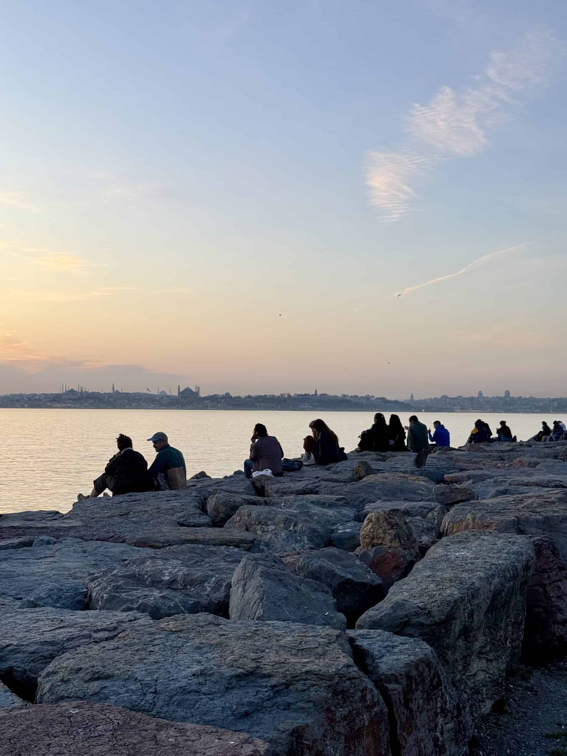 People sitting on the rocks of the Moda promenade looking out across the sea to the historical peninsula